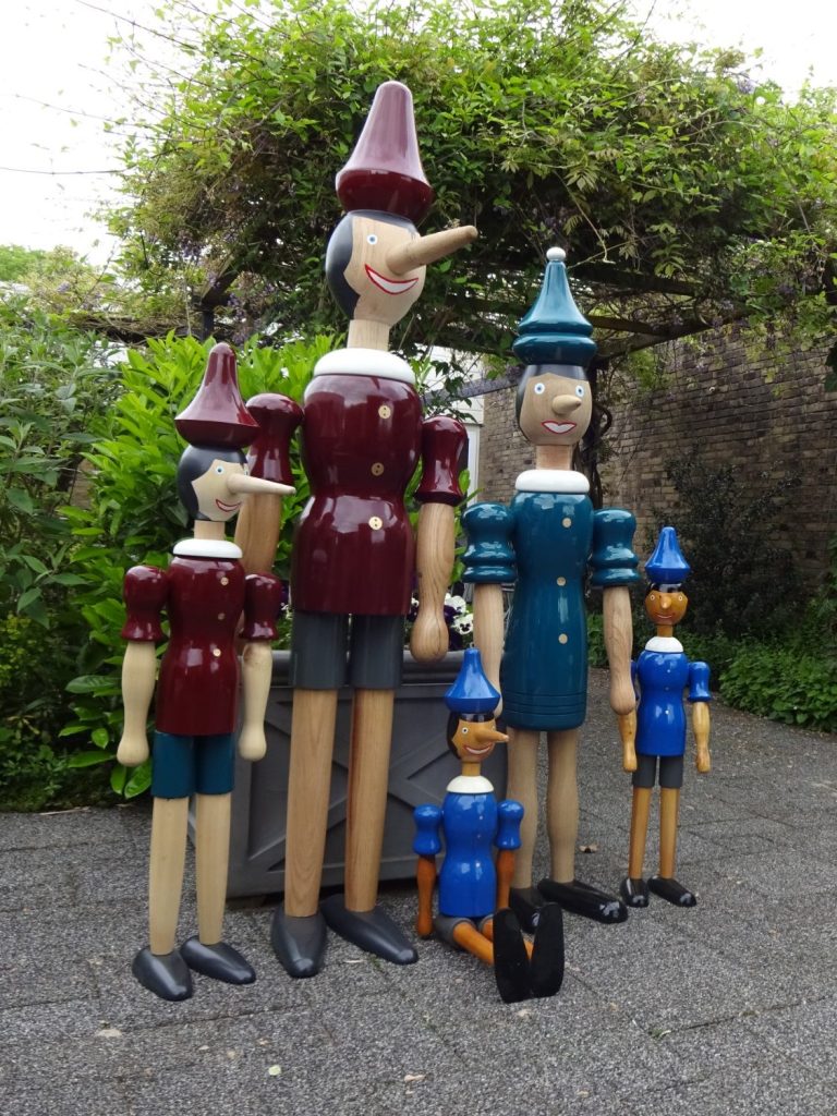 Vijf houten Pinocchio-figuren in verschillende groottes, met kleurrijke hoeden en kleding.